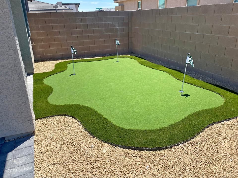 A small backyard putting green with three golf holes, featuring lush turf from a top-rated artificial grass installer in Apache Junction, AZ, is bordered by rocks and enclosed by a tan brick wall and neighboring houses.