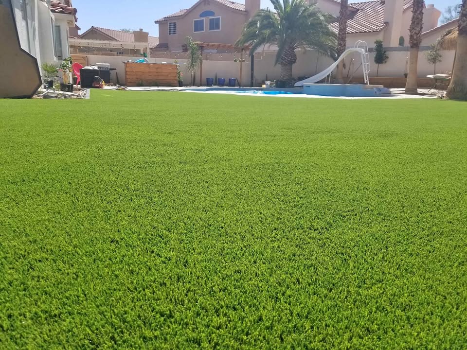 A backyard with bright green artificial grass installed by a top-rated artificial grass installer in Apache Junction, AZ, a swimming pool with a white slide, palm trees, and several houses in the background under a clear blue sky.