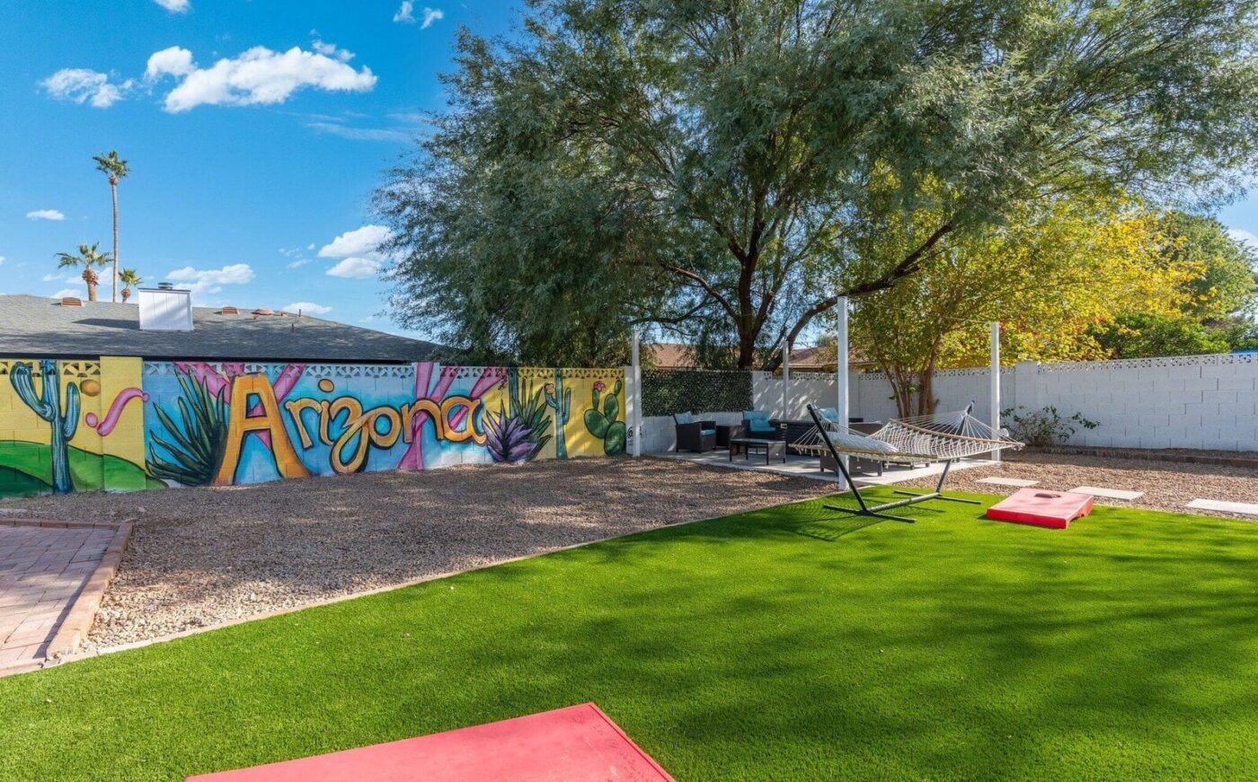 A backyard featuring top-rated artificial grass, cornhole boards, a hammock, outdoor seating, and a vibrant "Arizona" mural with cacti and desert scenery. Trees sway under a blue sky with clouds—installed by the best in Apache Junction, AZ.