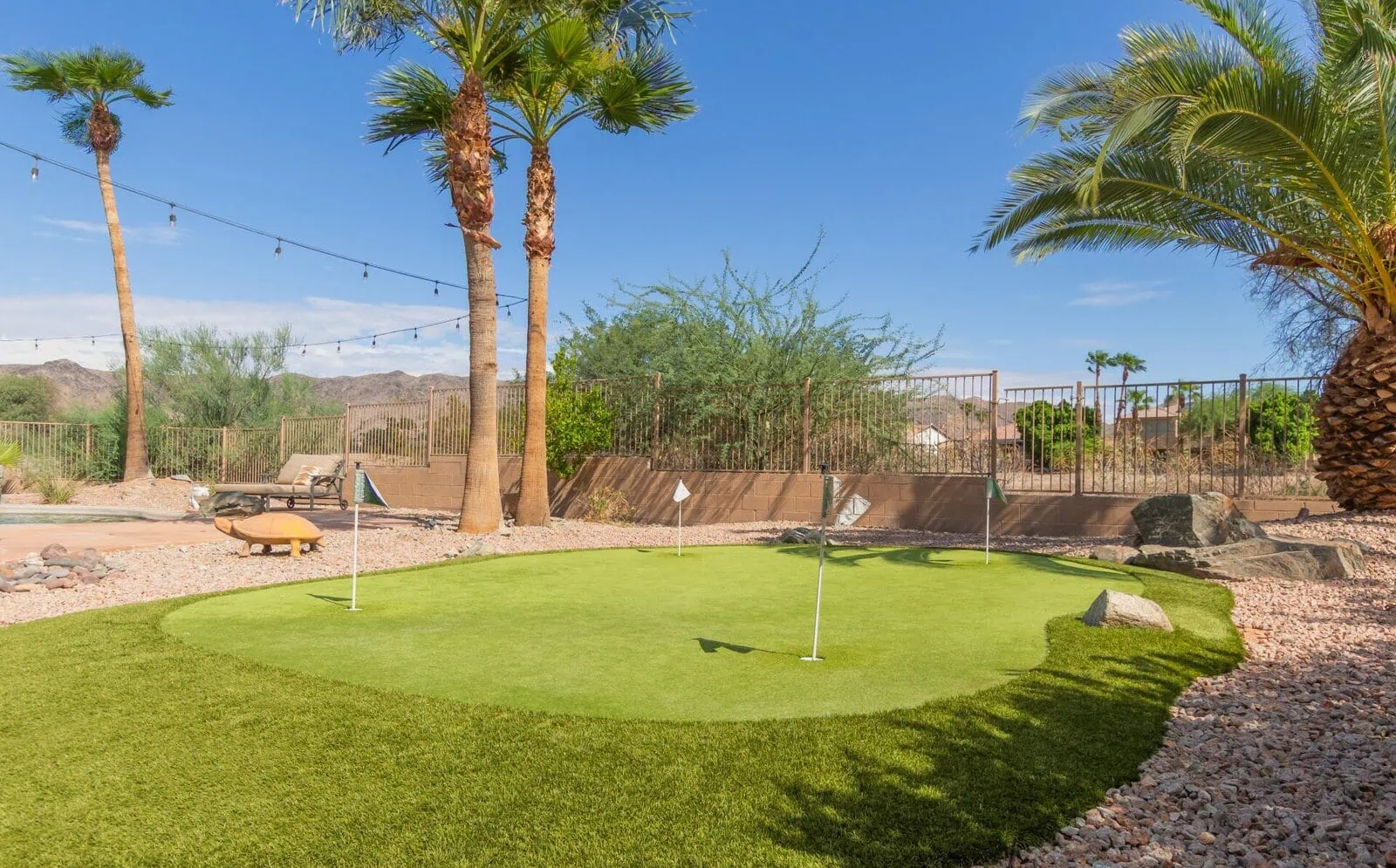A backyard putting green by a top-rated artificial grass installer in Apache Junction, AZ, with flags, rocks, palm trees, and desert plants, set against mountains under a clear blue sky. String lights hang between the trees.