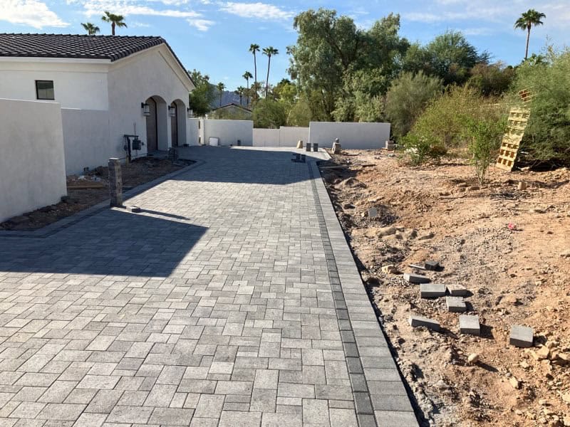 A newly installed gray paver driveway runs alongside a white house with an unfinished dirt area, ideal for a future lawn by a top-rated artificial grass installer in Apache Junction, AZ, with scattered pavers, trees, and a white wall in the background.