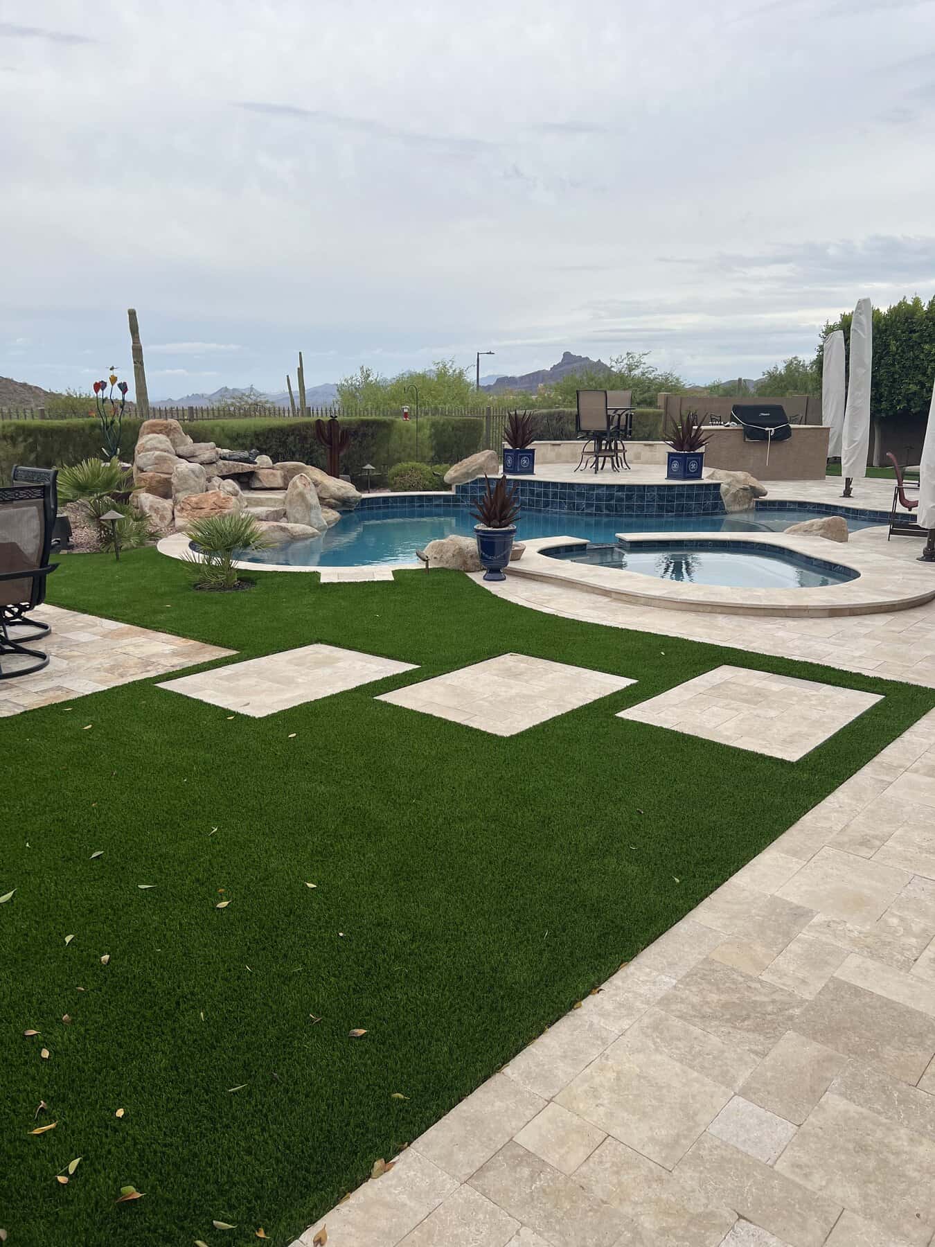 A backyard with a pool and hot tub surrounded by stone tiles and artificial grass, expertly installed by a top-rated artificial grass installer in Apache Junction, AZ. Lounge chairs, potted plants, and desert landscaping frame mountain views.