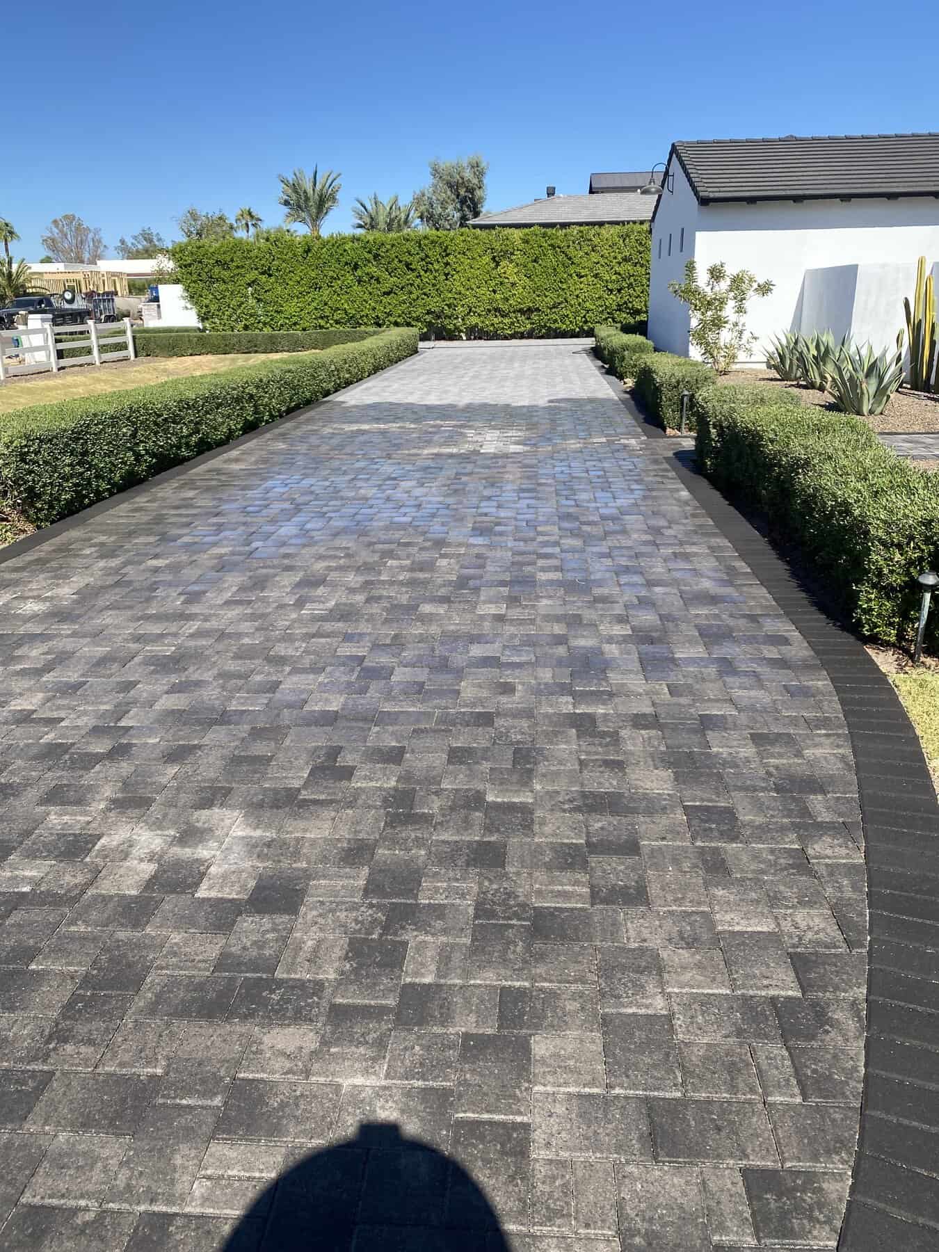 A paved driveway with a distinct light and dark pattern runs between green hedges—expertly installed by a top-rated artificial grass installer in Apache Junction, AZ—toward a white house with a black roof, under a clear blue sky.