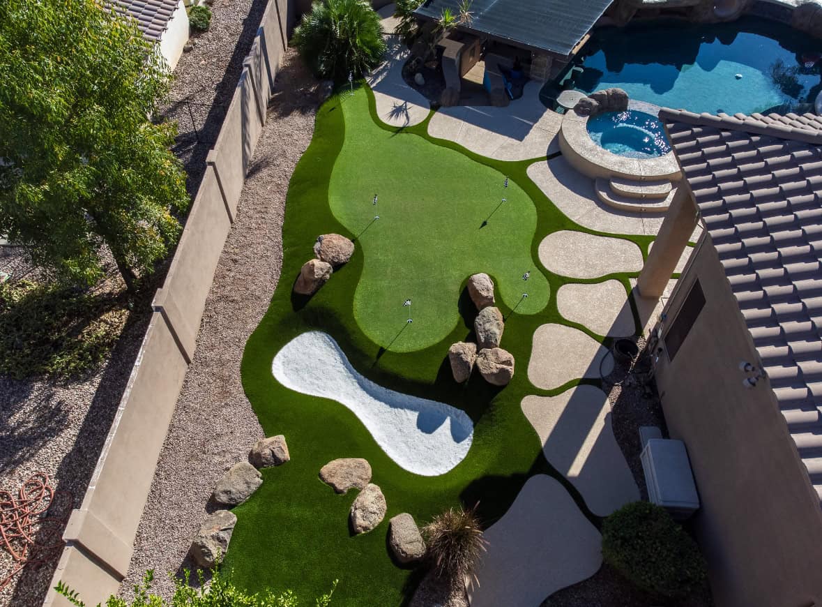 Aerial view of a backyard with a mini golf putting green, installed by a top-rated artificial grass installer in Apache Junction, AZ, featuring artificial turf, sand trap, large rocks, and a swimming pool surrounded by patio and landscaping.