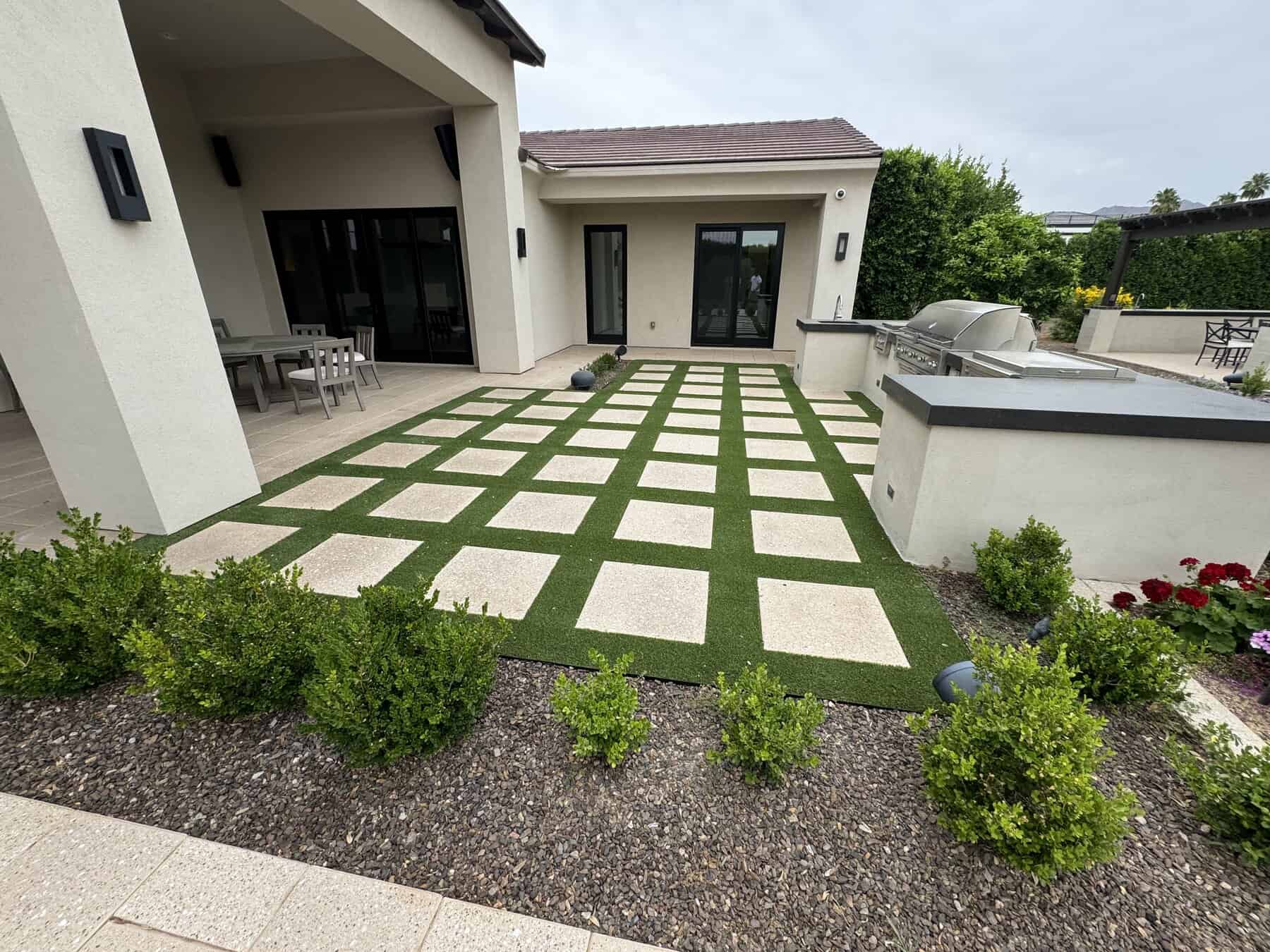 Modern backyard patio with a grid of square concrete pavers set in artificial grass by a top-rated artificial grass installer in Apache Junction, AZ, surrounded by shrubs and lush garden beds, next to an outdoor dining area and barbecue grill station.
