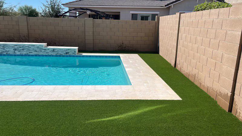 A backyard swimming pool with clear blue water is bordered by light-colored tiles and lush artificial grass, expertly installed by a Top-Rated Artificial Grass Installer in Apache Junction, AZ, and surrounded by tall beige brick walls under a sunny sky.