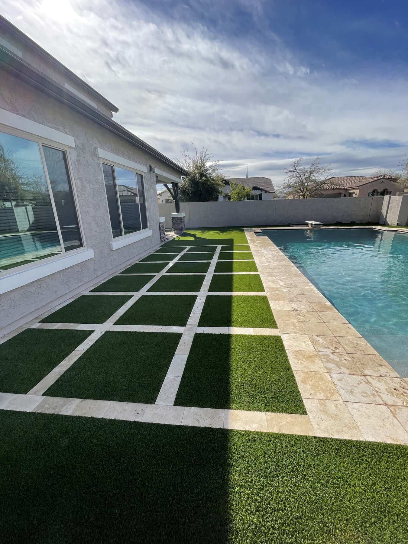 A backyard poolside area with a modern geometric pattern of alternating green grass and light stone tiles, designed by a top-rated artificial grass installer in Apache Junction, AZ, beside a house with large windows under a partly cloudy sky.