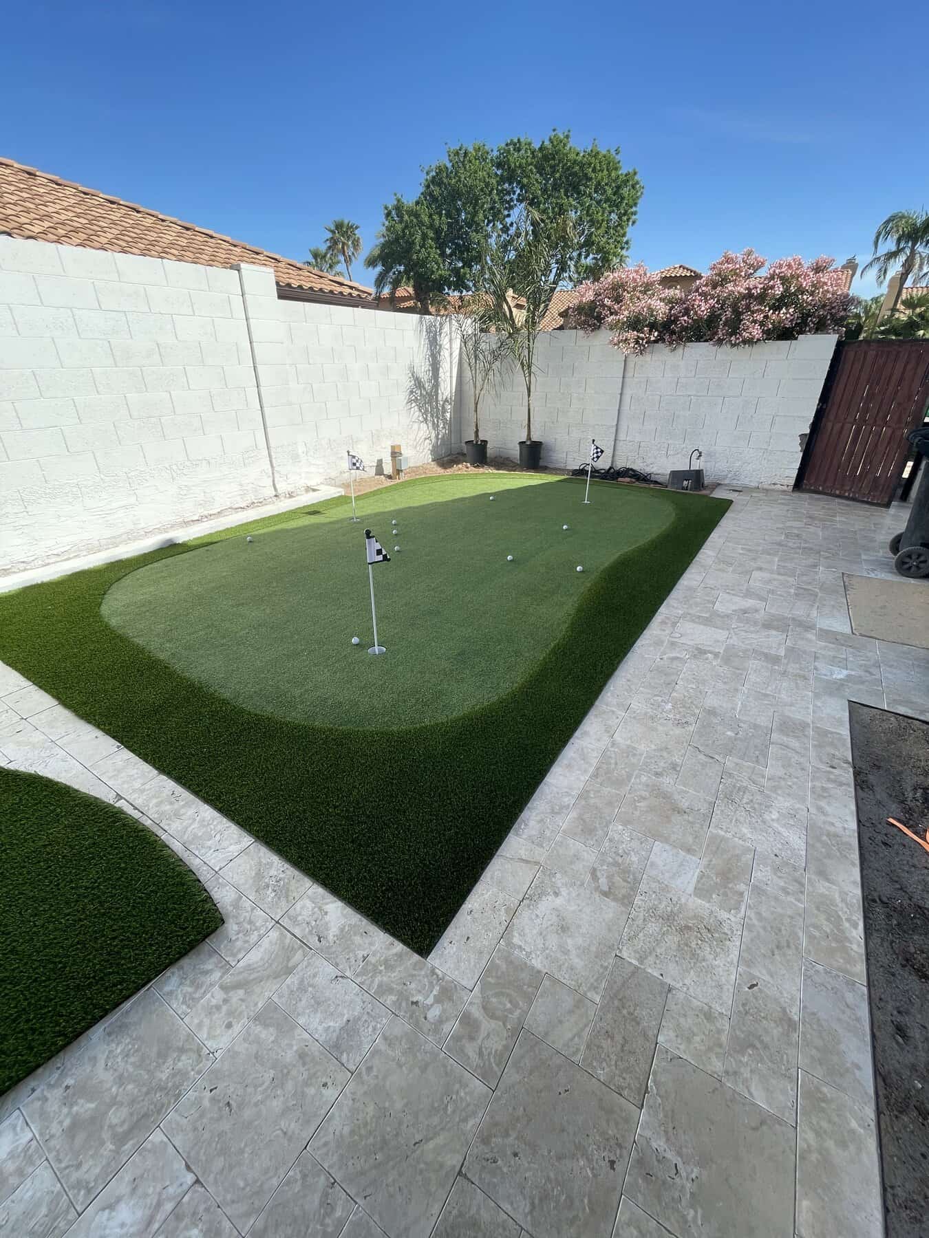 A backyard features a putting green with several golf holes, flags, and light-colored stone tiles. Installed by a top-rated artificial grass installer in Apache Junction, AZ, it’s framed by a white wall, green trees, and pink flowering shrubs under a clear blue sky.
