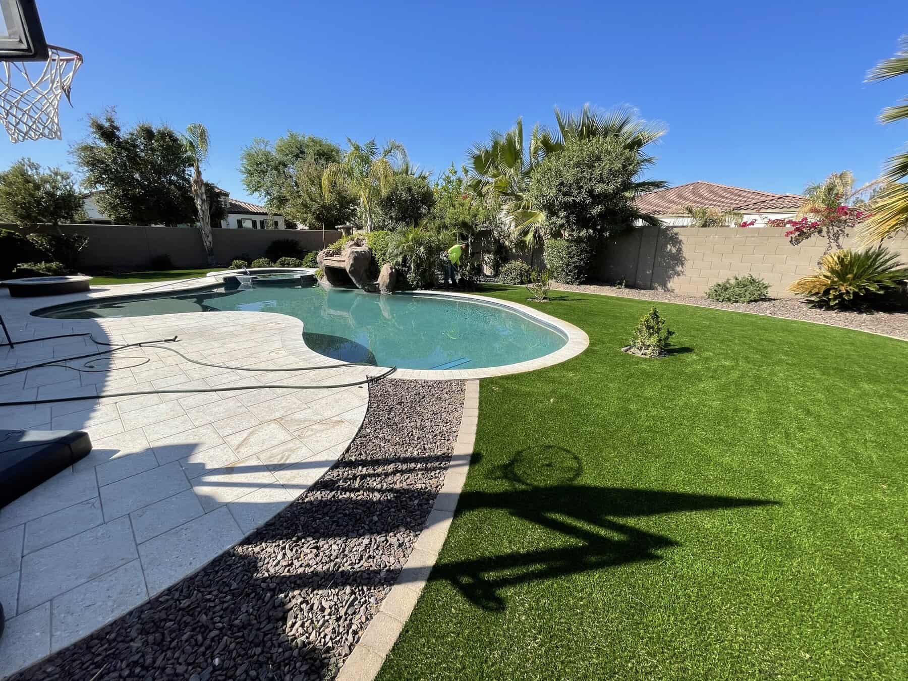 A backyard with a curving swimming pool is surrounded by palm trees, green artificial grass from a top-rated artificial grass installer in Apache Junction, AZ, paving stones, and a basketball hoop casting a shadow under a clear blue sky.