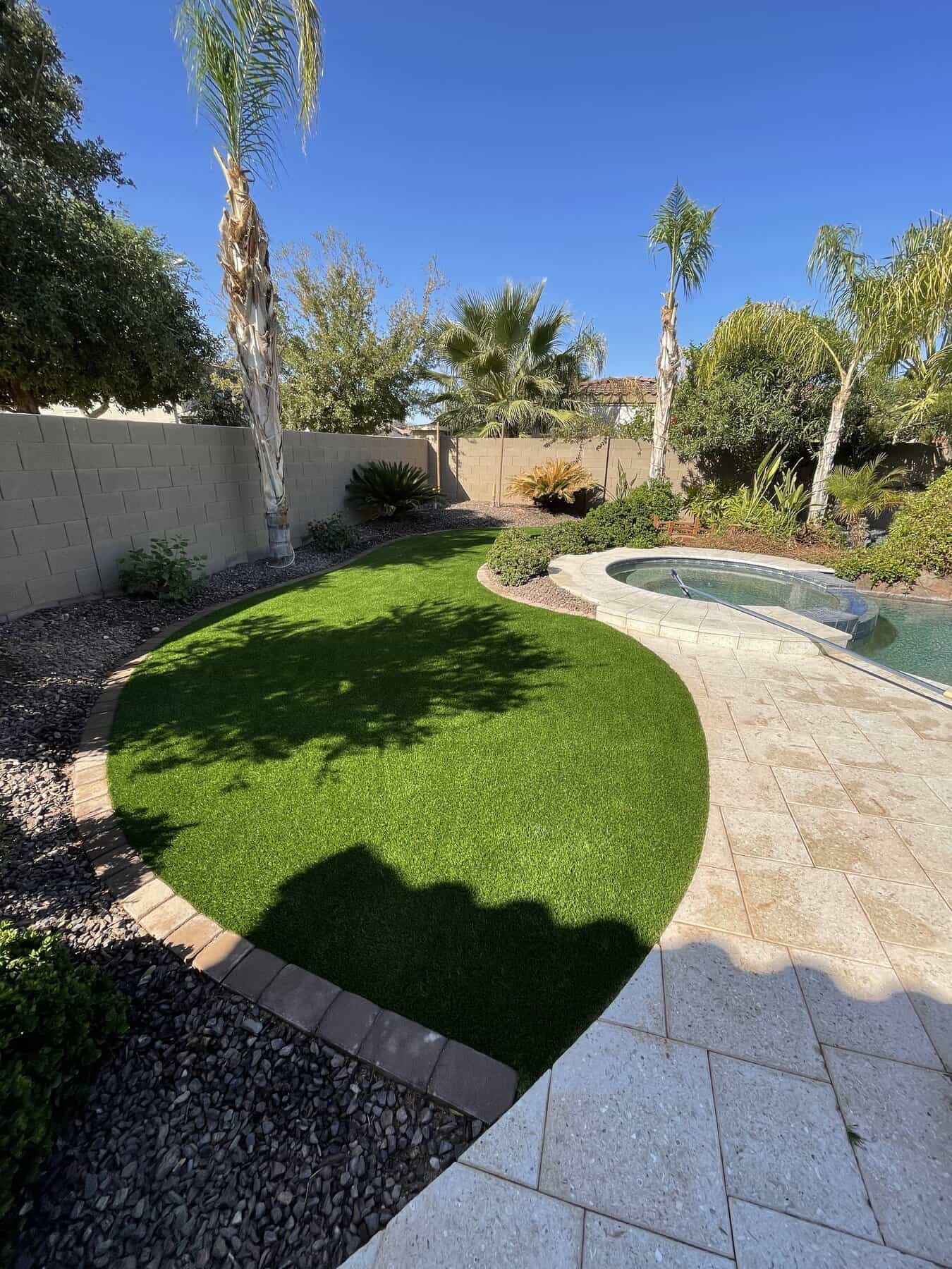 A backyard with a curved artificial grass lawn by a top-rated artificial grass installer in Apache Junction, AZ, bordered by tan bricks, a stone patio, a small pool or spa, palm trees, and shrubs under a clear blue sky.