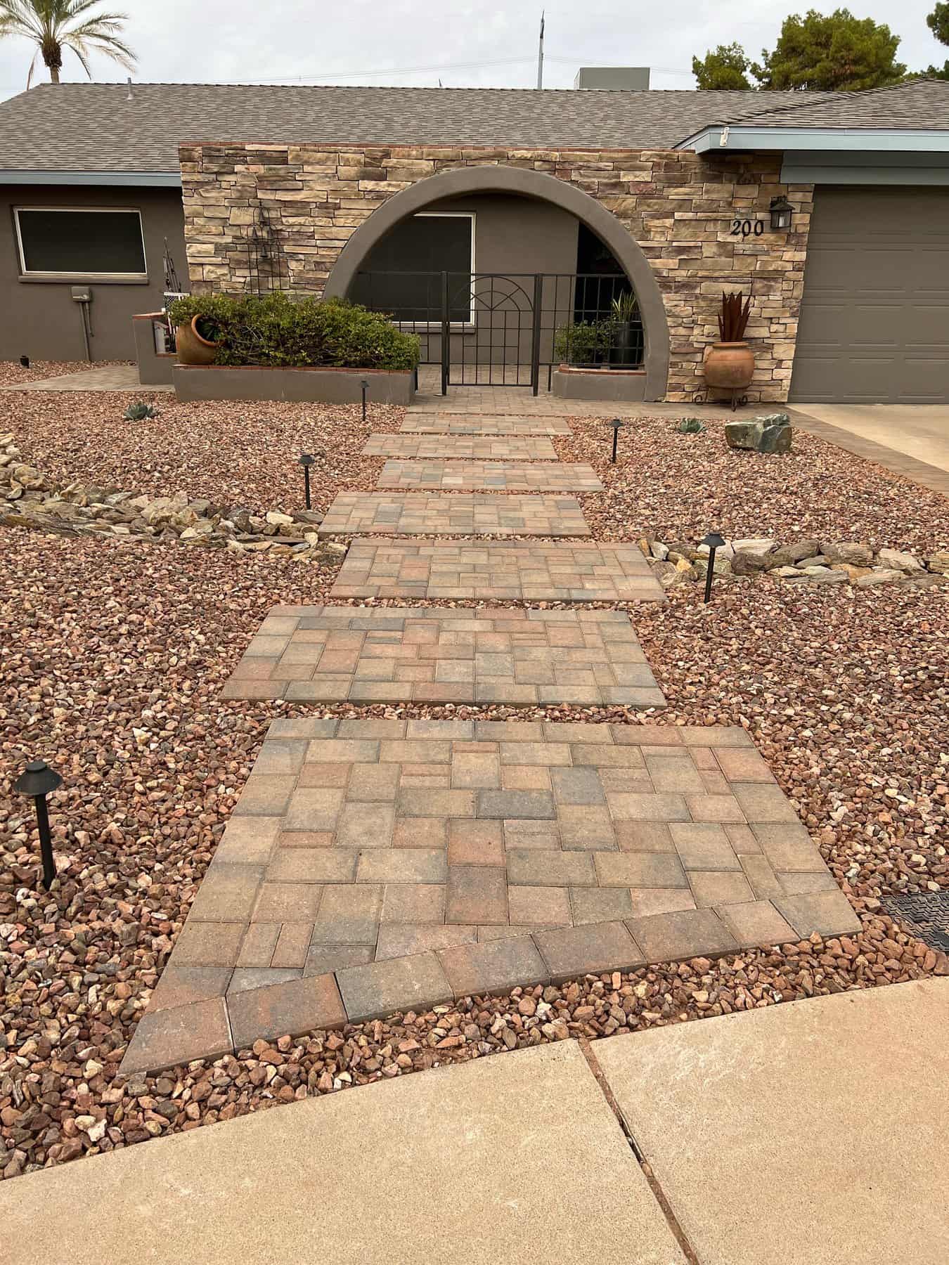 A stone walkway with square pavers winds through a front yard accented by brown rocks and top-rated artificial grass, leading to a house with a brick facade, arched entryway, and gray garage door. Small landscape lights line the path.