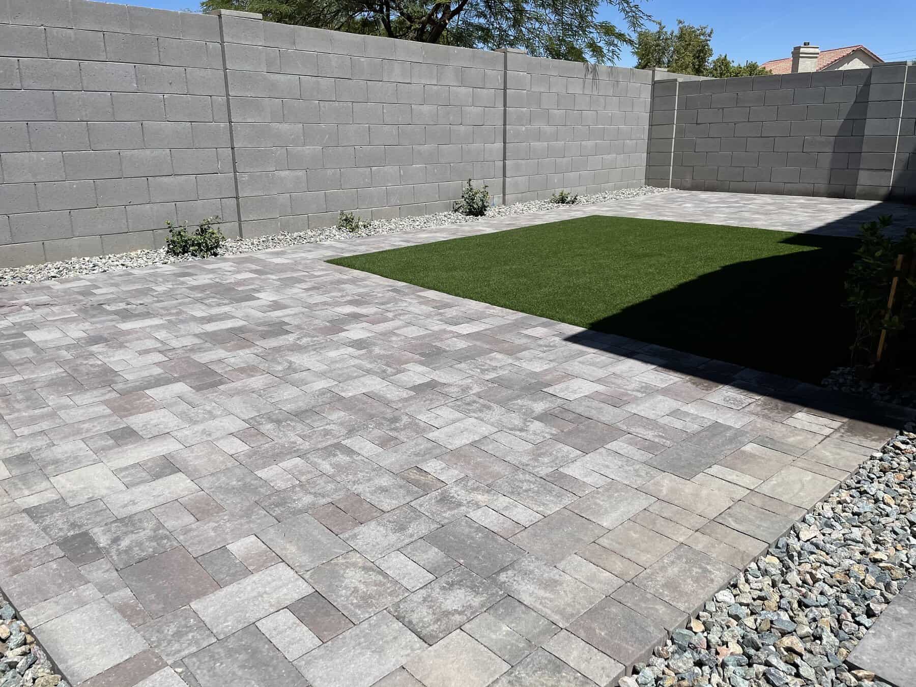 A backyard with gray stone pavers, a rectangular patch of artificial grass professionally installed by a top-rated artificial grass installer in Apache Junction, AZ, small plants along the block wall, and gravel borders enclosed by tall gray concrete walls.