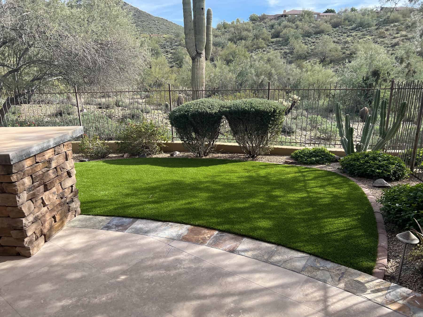 A backyard with a small patch of artificial grass, expertly installed by a top-rated artificial grass installer in Apache Junction, AZ, bordered by stone and desert plants, with mountains and a metal fence in the background.