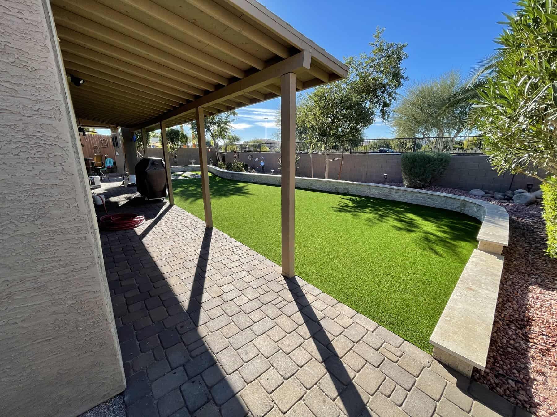 A backyard with a covered patio, paved walkway, artificial grass lawn installed by a top-rated artificial grass installer in Apache Junction, AZ, a curved stone bench, and landscaping with rocks and desert plants under a clear blue sky.