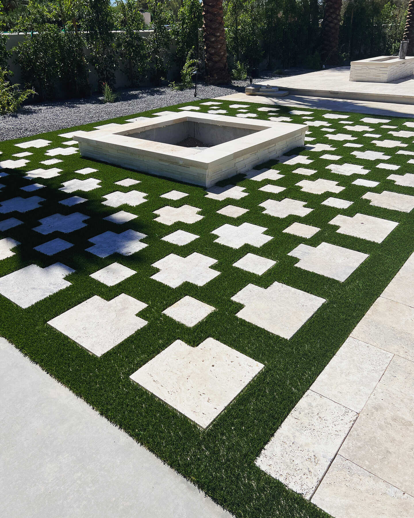 A modern outdoor patio features a square firepit surrounded by lush artificial grass, installed by a top-rated artificial grass installer in Apache Junction, AZ. Large, irregular stone tiles form a geometric pattern, bordered by trees and shrubs.