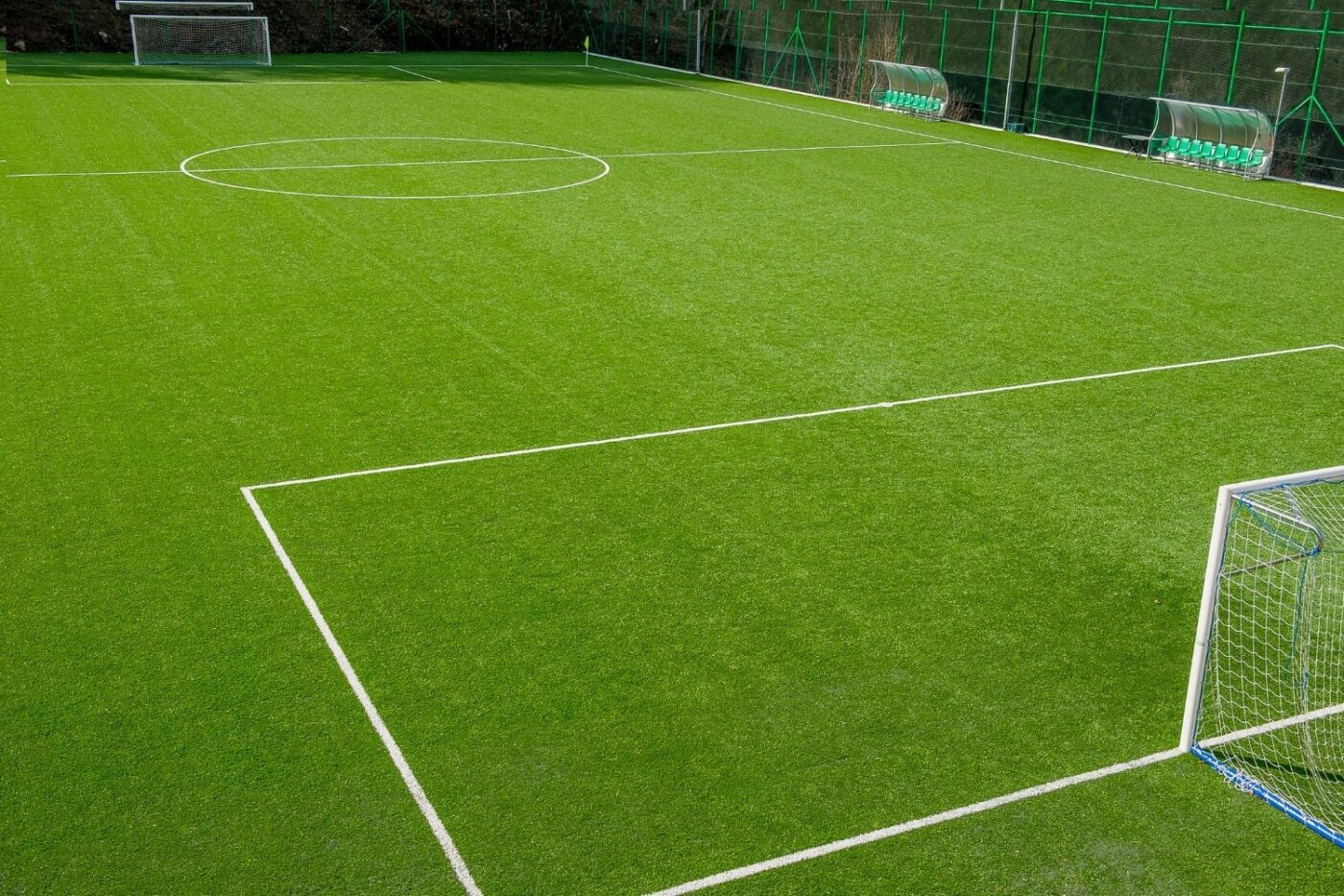 An empty outdoor sports field with bright green artificial turf, white boundary lines, two goals, and team benches along the side, all enclosed by a green fence.
