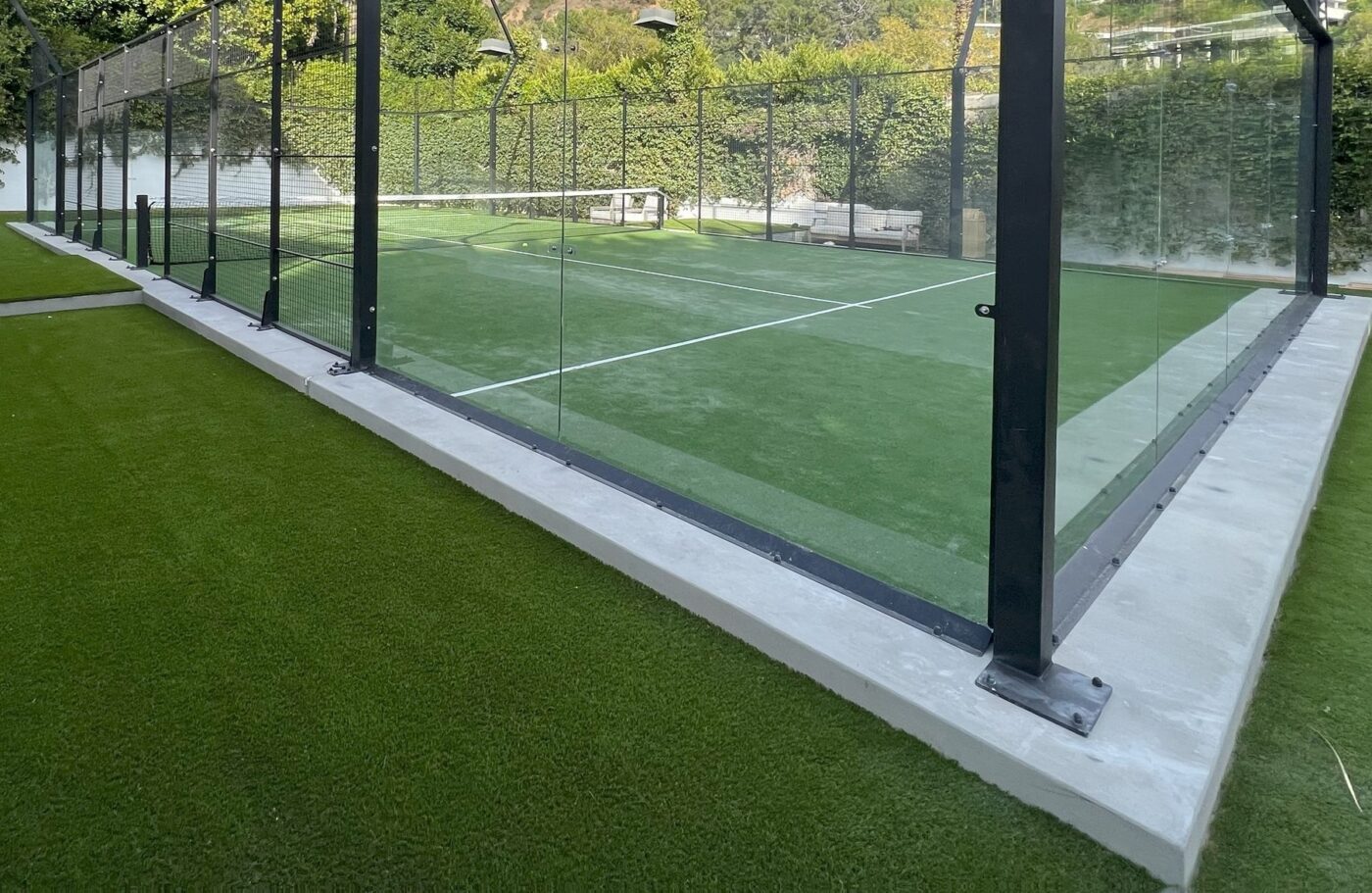 A fenced paddle tennis court with glass walls sits on vibrant sports field artificial turf, surrounded by trees. The empty court and outdoor setting create a peaceful atmosphere.