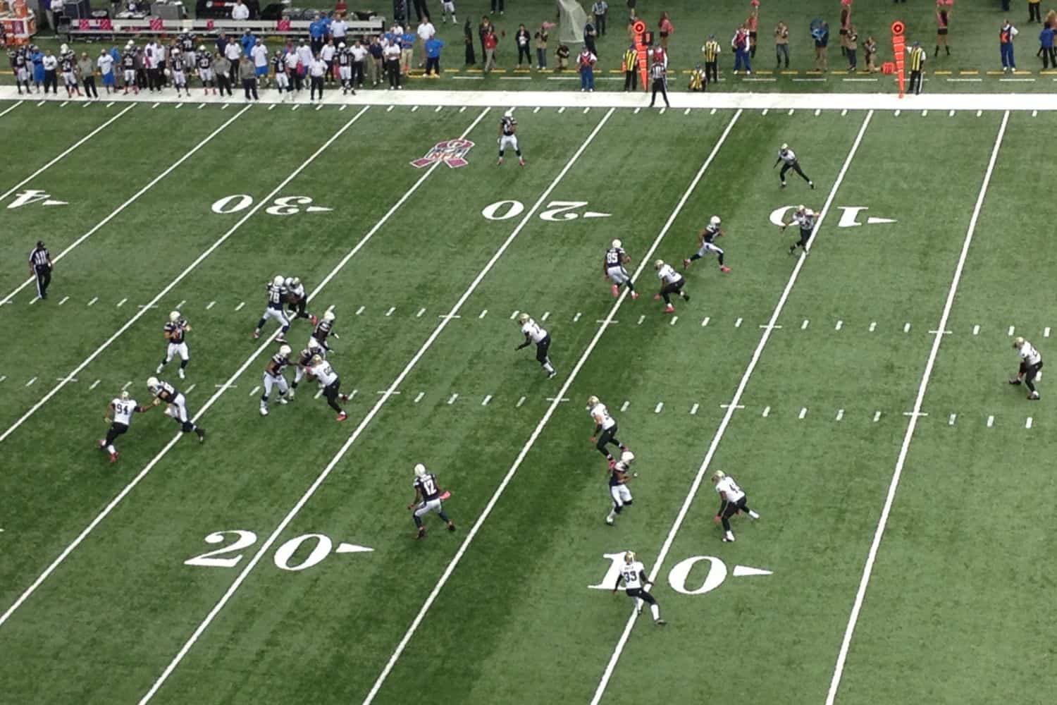 An aerial view of an American football game in progress on a sports field with artificial turf, where players in black and white uniforms spread out near the 20-yard and 10-yard lines as spectators watch from the stands.