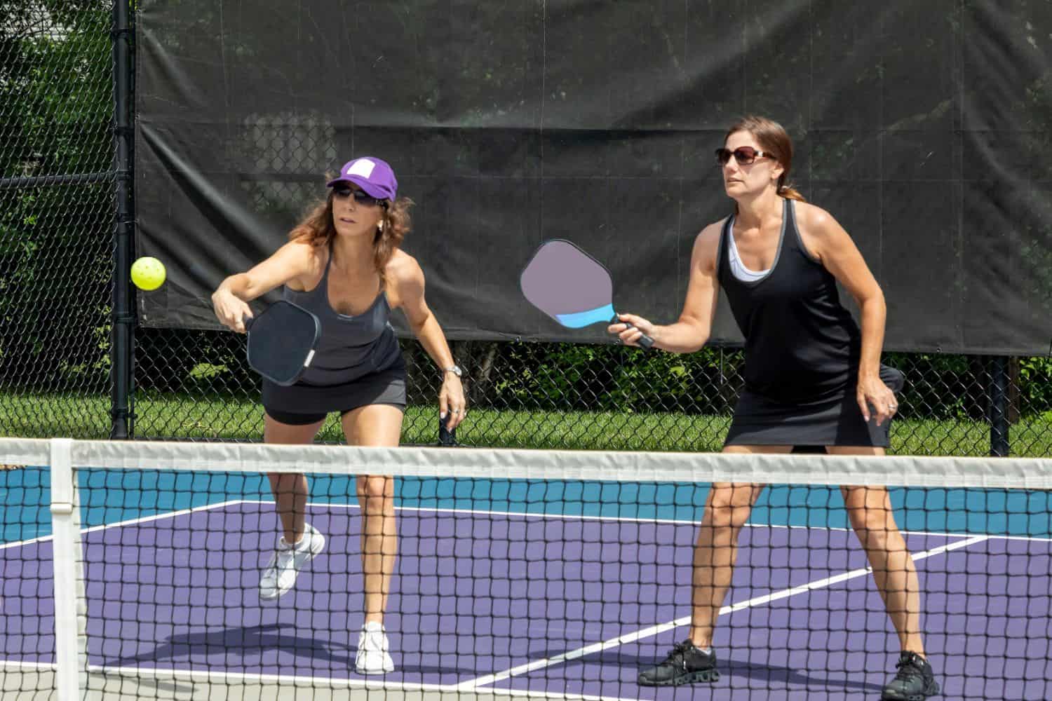 Two women playing pickleball on an outdoor sports field with artificial turf, both holding paddles and focused on a yellow ball in mid-air. One woman is preparing to hit the ball while the other stands ready.