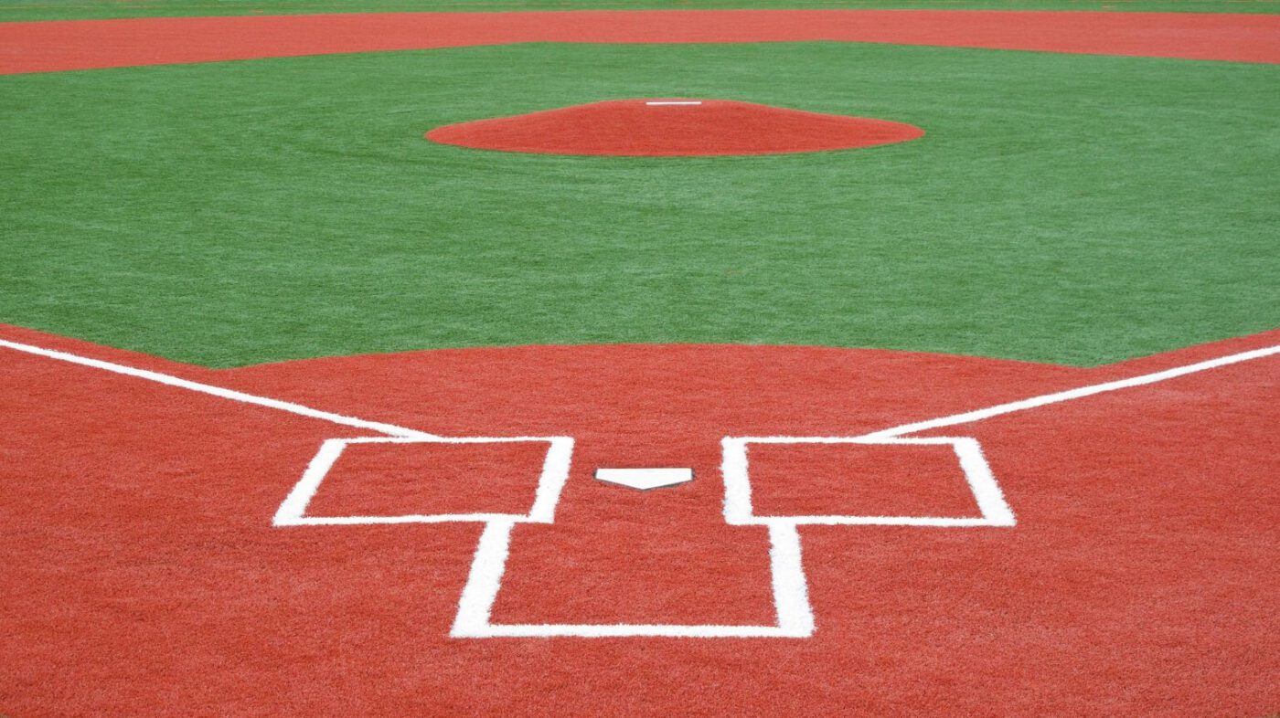 A view of a baseball field with Sports Field Artificial Turf, showing the home plate, batter’s boxes, catcher’s box, and pitcher’s mound. The green turf contrasts with the bright red infield and crisp white boundary lines.