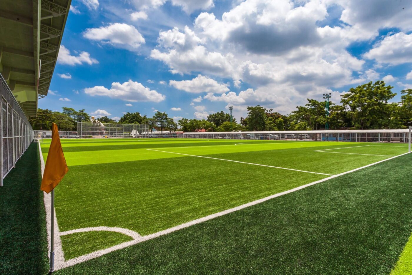 A bright, empty sports field with artificial turf, a yellow corner flag, and a partly cloudy blue sky. Trees and fencing surround the field, with a structure on the left side.