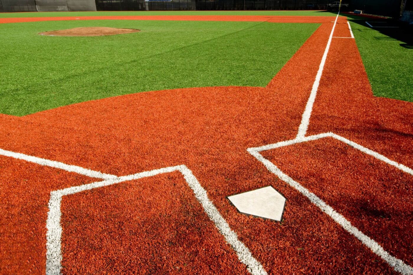 A close-up view of a sports field with bright artificial turf, showing home plate in the foreground and the pitcher's mound and base lines extending into the outfield.