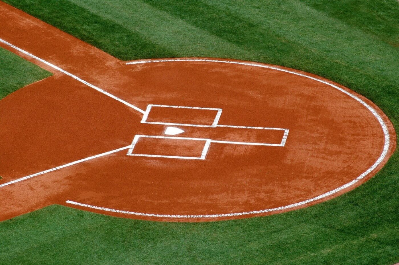 Aerial view of a baseball field’s home plate area, featuring reddish-brown dirt, white chalk lines marking batter’s boxes and base path, all surrounded by bright green Sports Field Artificial Turf.