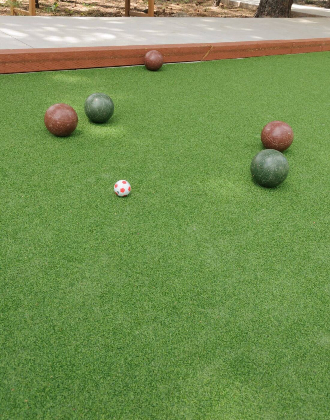 Four bocce balls, two green and two red, and a smaller white ball with red dots are positioned on an artificial turf sports field bocce court. A wooden border frames the court.