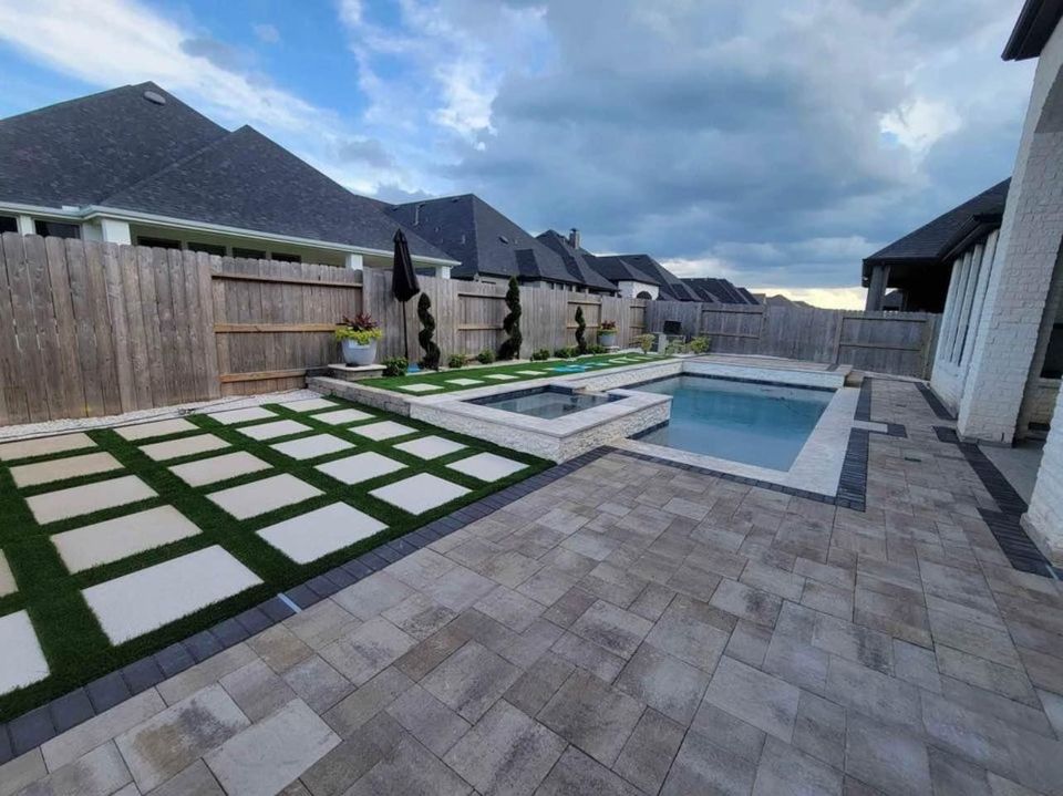 A modern backyard features a swimming pool with an attached hot tub, surrounded by light stone tiles. Pavers installation near me is evident in the patterned grass and paver area, planters, and wooden fencing beneath a cloudy sky.
